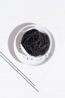 De cima de tigela de cerâmica com espaguete delicioso com tinta de lula preta com pauzinhos no fundo branco — Fotografia de Stock