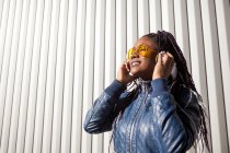 Щаслива молода афроамериканська самиця з африканськими плечима, одягнена в синю куртку і стилізовані сонцезахисні окуляри, яка насолоджується музикою через навушники і заливає сонячним світлом від смугастих стін. — стокове фото
