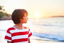 Вид збоку вдумливого хлопчика, що стоїть на мокрому піщаному березі, омитому махаючи блакитним морем на заході сонця — стокове фото