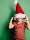 Garçon heureux méconnaissable couvrant visage avec chapeau rouge Santa debout sur fond vert — Photo de stock