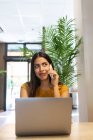 Mujer freelancer seria sentada en la mesa de madera en la cafetería y teniendo una llamada telefónica mientras escribe en netbook mirando hacia otro lado - foto de stock