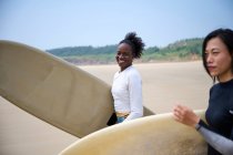 Посміхнена чорна спортсменка з довгим дошкою проти азіатської подруги з серфінгом, який дивиться вперед в океані під хмарним блакитним небом. — стокове фото