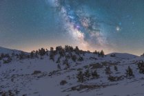 Paisagem de tirar o fôlego de encosta de colina coberta de neve e árvores contra altas montanhas rochosas sob céu estrelado de noite com Via Láctea — Fotografia de Stock