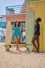Zufriedene junge multirassische Sportlerinnen mit Longboard und Surfbrett stehen am Sandstrand gegen Baumaßnahmen unter bewölktem Himmel — Stockfoto