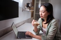 Seitenansicht einer jungen ethnischen Bloggerin am Schreibtisch mit Netbook und Kaffee, die im Hauszimmer in die Kamera schaut — Stockfoto