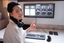 Lächelnde ethnische Maklerin tippt auf Tastatur gegen Monitor mit Grafik und zeigt wie Geste, während sie zu Hause in die Kamera schaut — Stockfoto