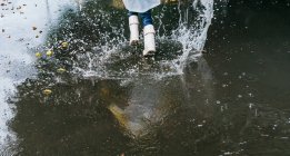 From above back view of crop anonymous child in rubber boots having fun in puddle with splattering aqua on rainy day — Stock Photo