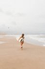 Visão traseira de esportista jovem irreconhecível em roupa de banho com prancha de surf olhando para longe na costa arenosa contra o oceano tempestuoso — Fotografia de Stock