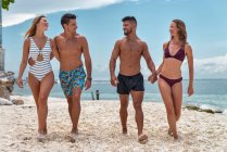 Cheerful tourists in swimwear talking while strolling on sandy shore against ocean under cloudy blue sky in town — Stock Photo