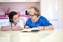 Abuela concentrada en ropa casual y anteojos sentados a la mesa y leyendo libro con nieta alegre en la cocina en casa - foto de stock