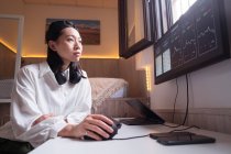 Сторона зору концентрованої азіатської жінки, що працює на комп'ютері з графіками, що показують динаміку змін в вартості криптовалют на зручному робочому місці. — стокове фото