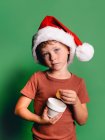 Adorable petit garçon avec chapeau de Noël Père Noël prendre cookie de tasse sur fond vert en regardant la caméra — Photo de stock