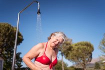 Fröhliche Seniorin im Bikini genießt Spritzer von der Dusche in der Nähe des Pools mit klarem Wasser — Stockfoto