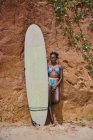Front view of African American female athlete looking at camera with surfboard from an area of the beach and in front of a clay rock with plants on its side — Stock Photo