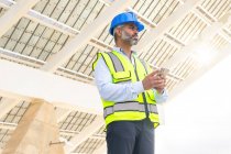 From below middle aged Hispanic foreman in in hardhat and waistcoat browsing on smartphone looking away while standing near solar power station — Stock Photo