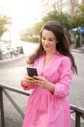 Улыбающаяся женщина в платье, стоящая на тротуаре и смс-ки на телефоне — стоковое фото