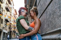 Vista lateral da moda alegre jovem casal lésbico com tatuagem em óculos de sol abraçando olhando um para o outro no momento do beijo encostado em uma parede na cidade — Fotografia de Stock