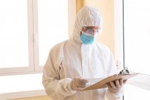 Adult male medic in personal protective equipment reading paper on clipboard during COVID 19 pandemic in hospital — Stock Photo