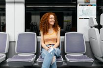 Содержание молодой женщины в разорванных джинсах с кудрявыми рыжими волосами, смотрящей на сиденье во время поездки на поезде — стоковое фото