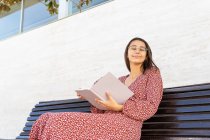 Positive young female in stylish clothes sitting with opened book on wooden bench against building with light wall in daytime with eyes closed — Stock Photo