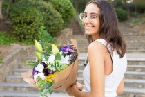 Contenida joven hembra en gafas de vista mirando a la cámara de pie con flores florecientes ramo de flores en las escaleras urbanas - foto de stock