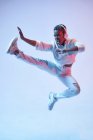 Энергичная этническая женщина в беспроводных наушниках и модной одежде прыгает с поднятой ногой и открытым ртом во время танца хип-хопа — стоковое фото