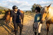 Smiling female and male trainer in protective helmets strolling with stallions on sandy terrain under cloudy sky — Stock Photo