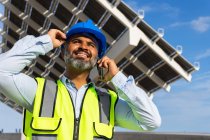 From below happy middle aged Hispanic foreman in in hardhat and waistcoat speaking on smartphone looking away while standing near solar power station — Stock Photo