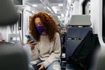 Увага жінка з кучерявим волоссям в масці для обличчя серфінг Інтернет на мобільному телефоні під час подорожі поїздом — стокове фото
