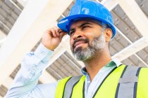 From below of mature ethnic male supervisor in vest putting on hardhat while looking away standing near solar power station — Stock Photo