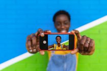 Concentration sélective du téléphone portable dans les mains d'une femme afro-américaine joyeuse prenant autoportrait contre un mur lumineux — Photo de stock