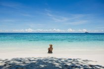 Voltar vista turista feminino em maiô e chapéu de palha sentado em mar transparente durante a viagem na Malásia — Fotografia de Stock