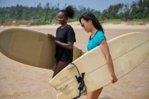 Lächelnde schwarze Sportlerin mit Longboard gegen asiatische Freundin mit Surfbrett, die sich im Meer unter wolkenlosem blauen Himmel freut — Stockfoto