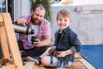 Hipster dad pouring herbal tea from thermos into calabash gourd against cheerful boy with lumber on boardwalk — Stock Photo
