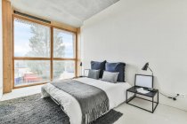 Design creativo della camera da letto con cuscini su piumino sul letto tra tavolo con lampada e finestra in casa — Foto stock
