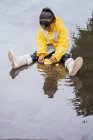 Очаровательный этнический ребенок в плаще играет с пластиковыми утками, отражающимися в волнистой луже в дождливую погоду — стоковое фото