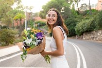 Vue latérale de la jeune femme amicale dans des lunettes avec bouquet floral en fleurs regardant loin sur la route de la ville — Photo de stock
