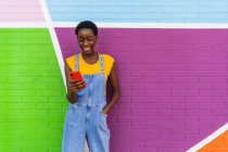 Mujer afroamericana en overoles de mezclilla de pie cerca de la pared colorida y el teléfono celular de navegación - foto de stock