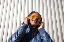 Щаслива молода афроамериканська самиця з африканськими плечима, одягнена в синю куртку і стилізовані сонцезахисні окуляри, яка насолоджується музикою через навушники і заливає сонячним світлом від смугастих стін. — стокове фото