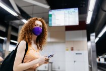 Вид збоку жінки в текстильній масці з мобільним телефоном і рюкзаком, дивлячись на платформу метро — стокове фото