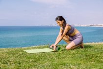 Contenu jeune asiatique femelle en vêtements de sport déroulant tapis de yoga sur la côte de l'herbe contre l'océan sans fin dans la lumière du soleil — Photo de stock