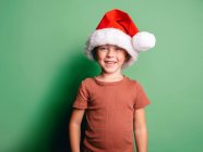 Positivo niño pequeño con el sombrero de Santa rojo sonriendo ampliamente contra el fondo verde y mirando a la cámara - foto de stock
