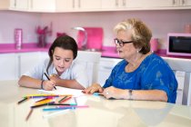 Улыбающаяся бабушка помогает веселой внучке рисовать на бумаге, проводя время вместе на кухне дома — стоковое фото