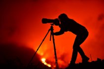 Vista laterale della silhouette di una donna accovacciata che fotografa con un teleobiettivo e treppiede l'esplosione di lava a La Palma Isole Canarie 2021 — Foto stock
