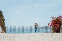 Ganzkörper junge Frau trägt trendige Jeans und Top steht auf dem Dach eines Hauses an der Küste des blauen Meeres — Stockfoto