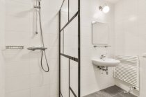 Minimalistisches Design des weißen Badezimmers mit Waschbecken unter Spiegel, das an der gefliesten Wand in der Nähe der Glasduschkabine in der Wohnung hängt — Stockfoto