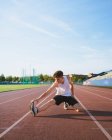 Fit male athlete in sportswear leaning forward while squatting during workout on track under cloudy sky in sunlight — Stock Photo