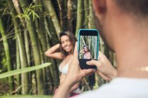 Анонімний чоловік, який подорожує врожаєм, фотографує усміхнену кохану жінку на мобільному телефоні проти бамбукових рослин у денний час — стокове фото