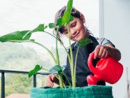 Focused little girl in black apron standing and watering green plant in pot on balcony against green hill in daytime — Stock Photo