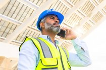From below middle aged Hispanic foreman in in hardhat and waistcoat speaking on smartphone looking away while standing near solar power station — Stock Photo